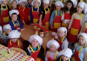 Cała grupa dzieci ubrana w stroje kucharzy pozuje do zdjęcia obok stolika, na którym stoją gotowe słoiki do zagotowania w kuchni.